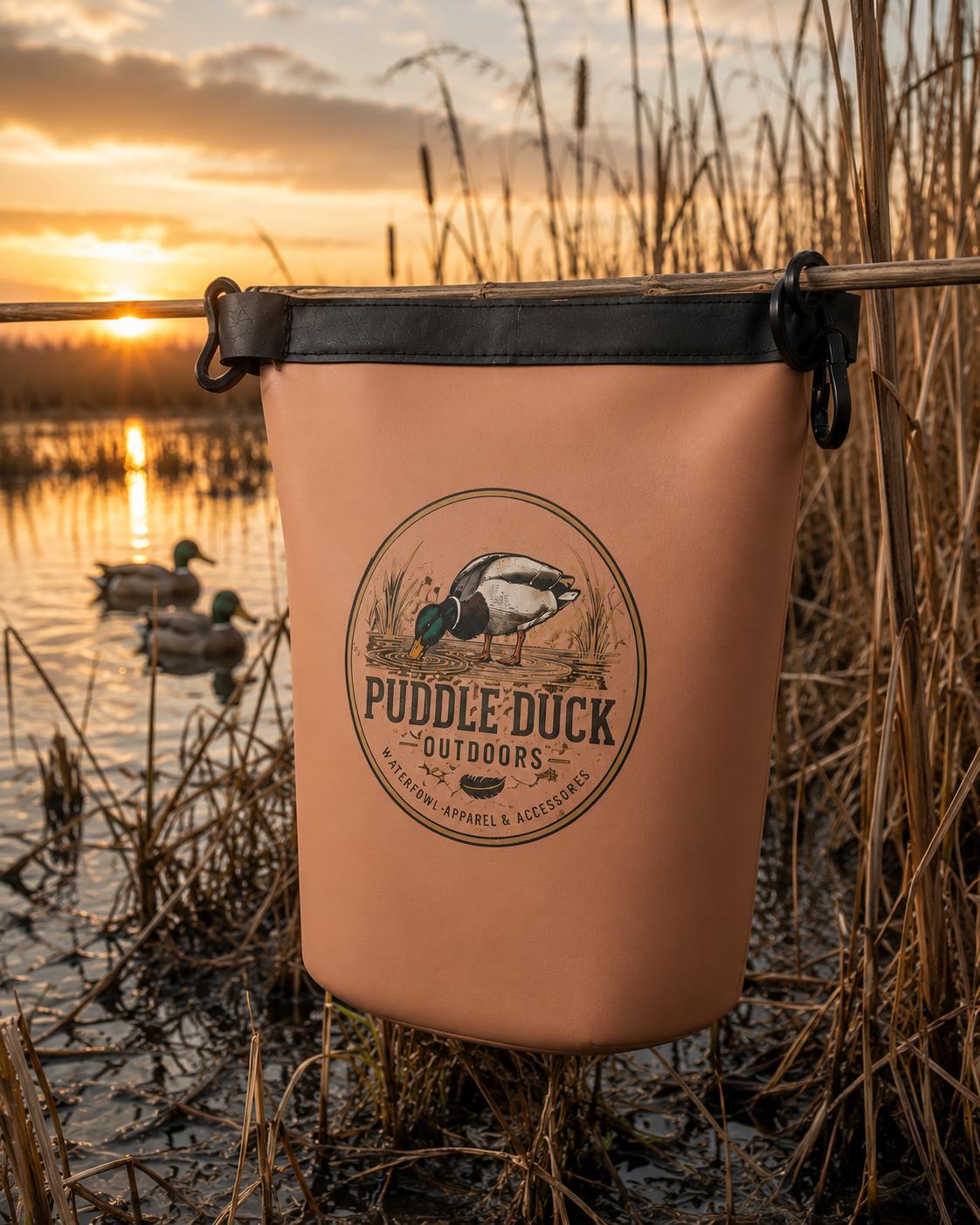A peach-colored duck blind or bag with a black top trim and a circular 'Puddle Duck Outdoors' logo, hanging from a rod over a marsh at sunset with ducks in the water.