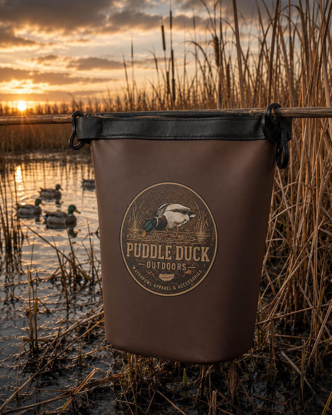 A brown bag with a black top trim and a circular 'Puddle Duck Outdoors' logo, hanging from a branch in a marshy wetland at sunset with ducks in the background.