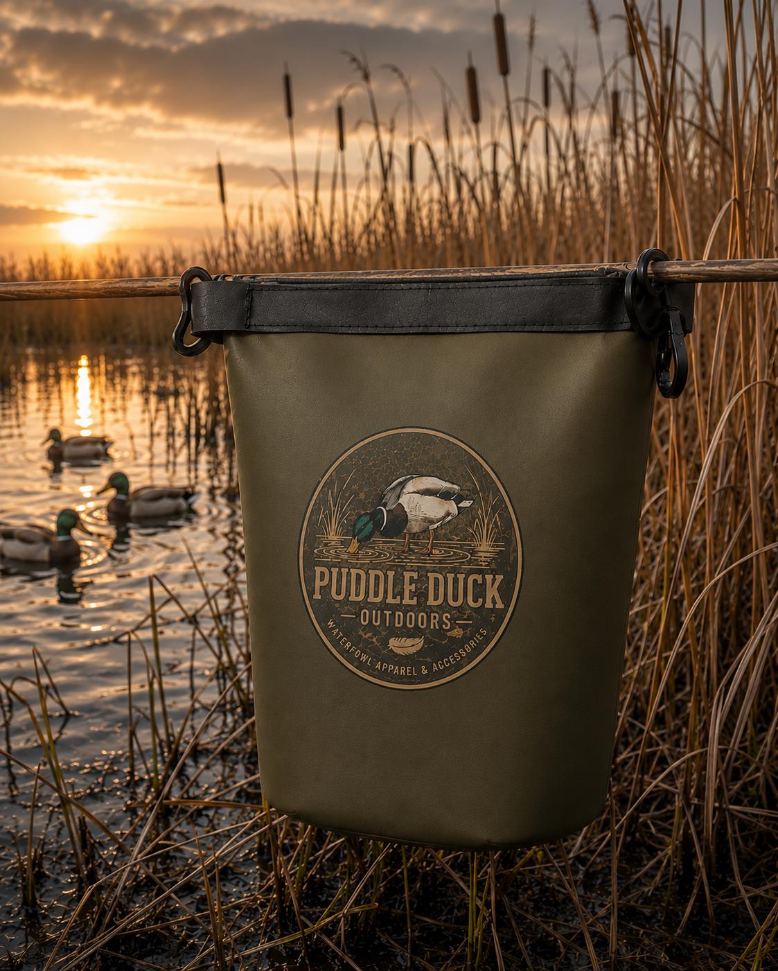 An olive green waterproof bag with a black top strap and a circular 'Puddle Duck Outdoors' logo, hanging from a wooden rod in a marshy wetland at sunset with ducks in the background.