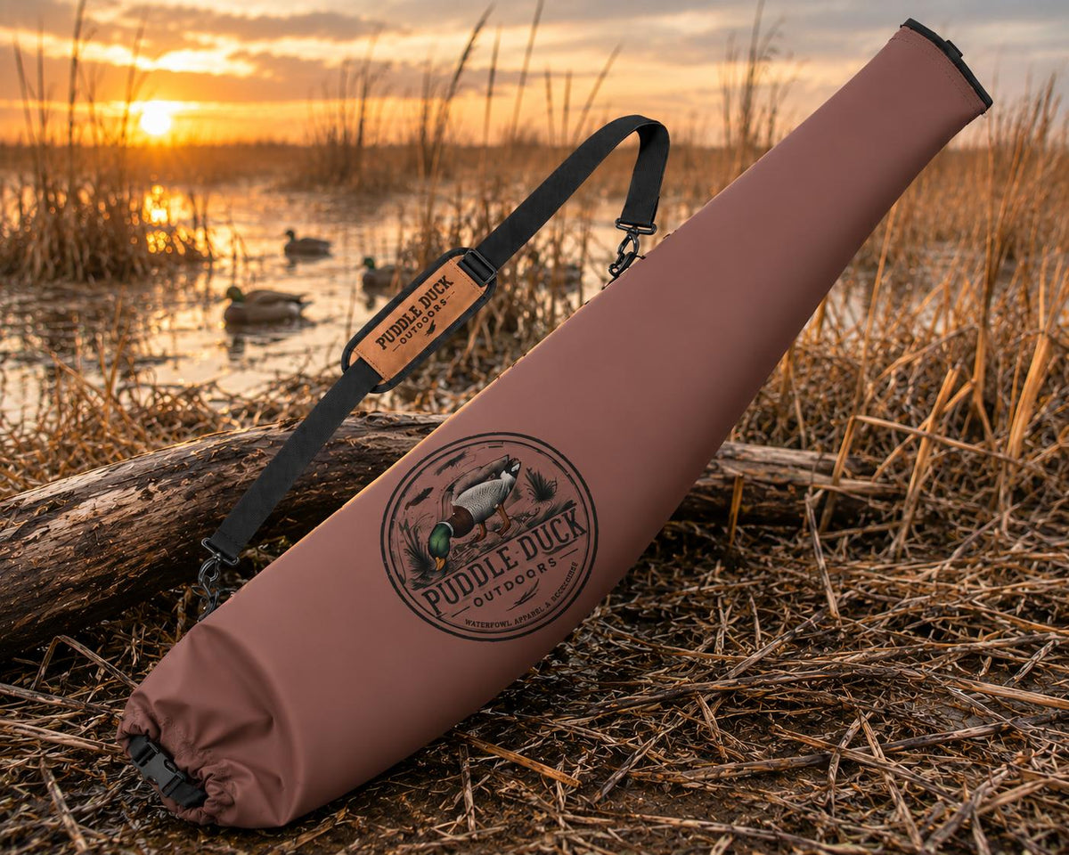 A duck hunting quiver bag in dusty rose pink lies on a log in a marsh at sunset. The bag features a black shoulder strap with a tan leather patch reading 'Puddle Duck Outdoors' and a circular logo on the front.