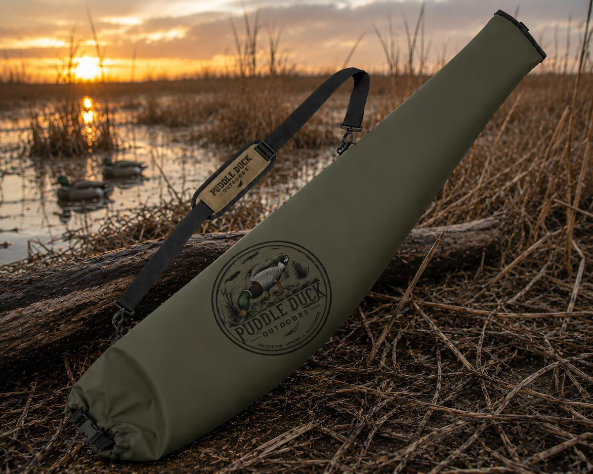An olive green Puddle Duck Outfitters bow case with a black shoulder strap, resting on a log in a marsh during a golden sunset.