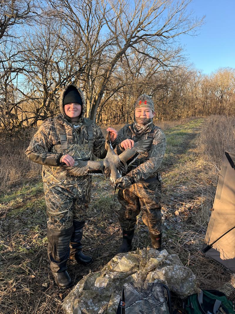 Two people in camouflage hunting suits standing outdoors in a wooded area, holding a harvested pintail. A camouflage tent is visible on the right, and bare trees and blue sky are in the background.