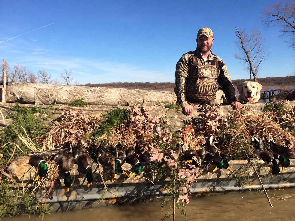 A man in camouflage hunting attire stands on a boat with a large haul of duck hunting deco bundles and ducks, with a dog beside him, outdoors under a blue sky.