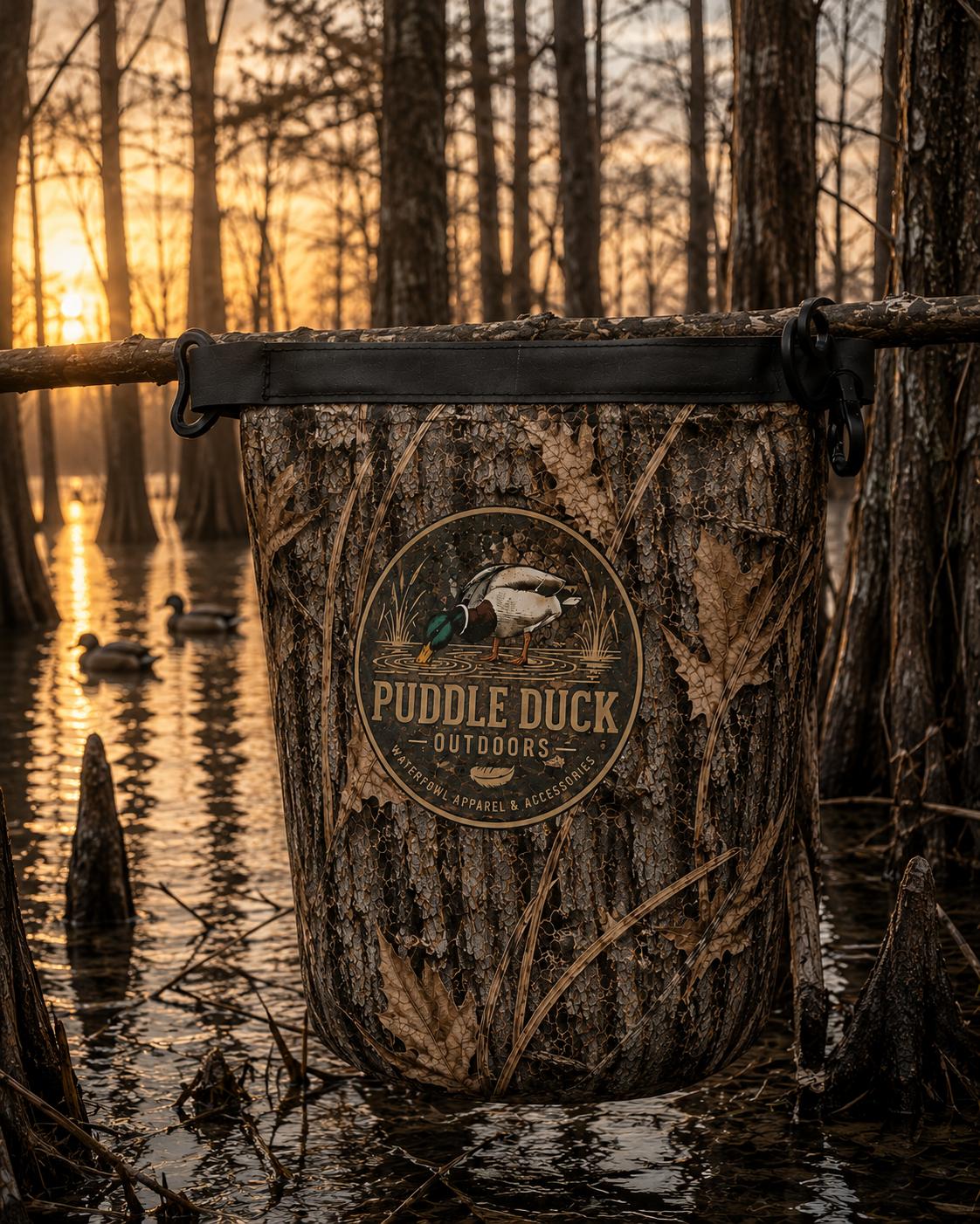 A camouflage 2 Liter waterproof bag hanging from a tree branch in a swamp at sunset, with a circular 'Puddle Duck Outdoors' logo on the front.