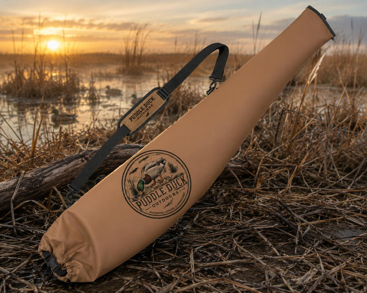 A tan Puddle Duck Outfitters shotgun case with a black shoulder strap, resting on a marsh grassy bank at sunset.