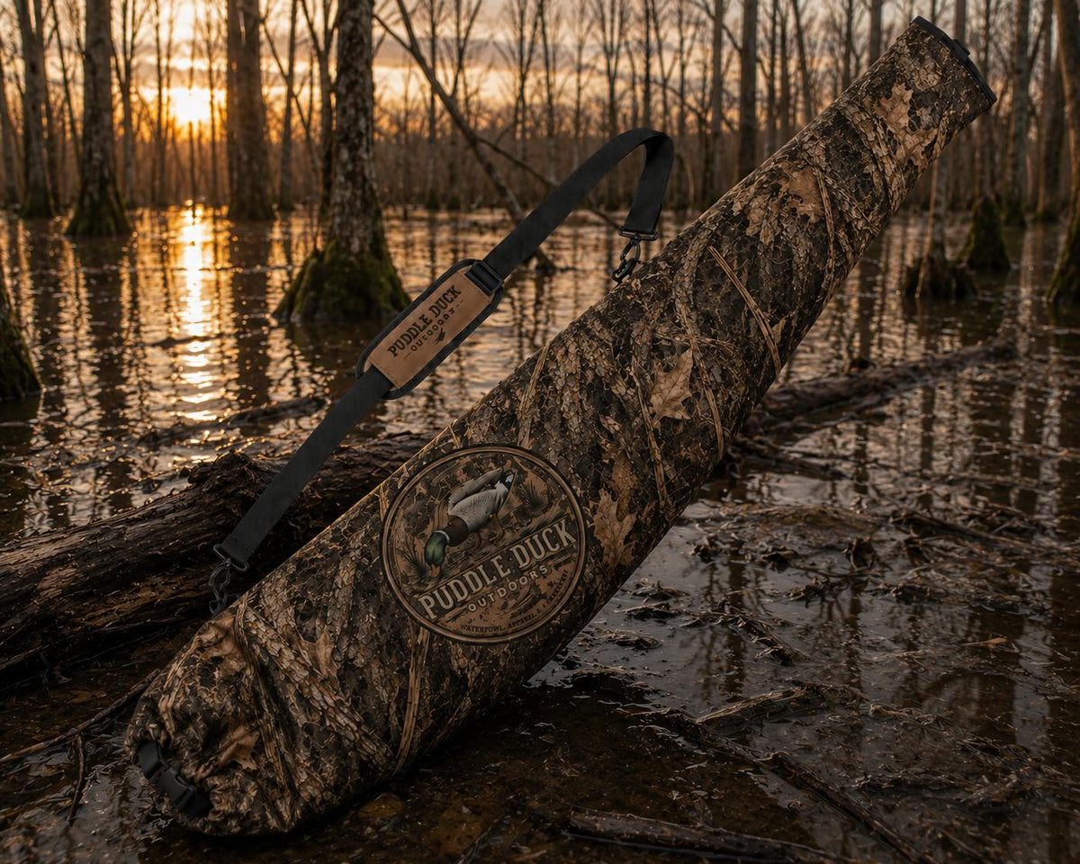 A camouflage duck hunting case with a black shoulder strap rests on a log in a swampy forest at sunset. The case features a circular 'Puddle Duck Hunting' logo patch.