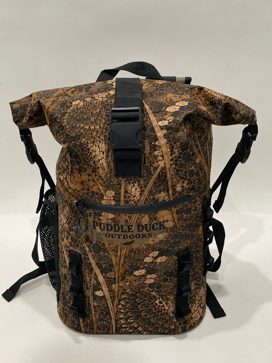 A roll-top backpack with a brown and beige marsh grass camouflage pattern, featuring a black plastic buckle strap on the front and the 'PUDDLE DUCK OUTDOORS' logo on the front pocket.